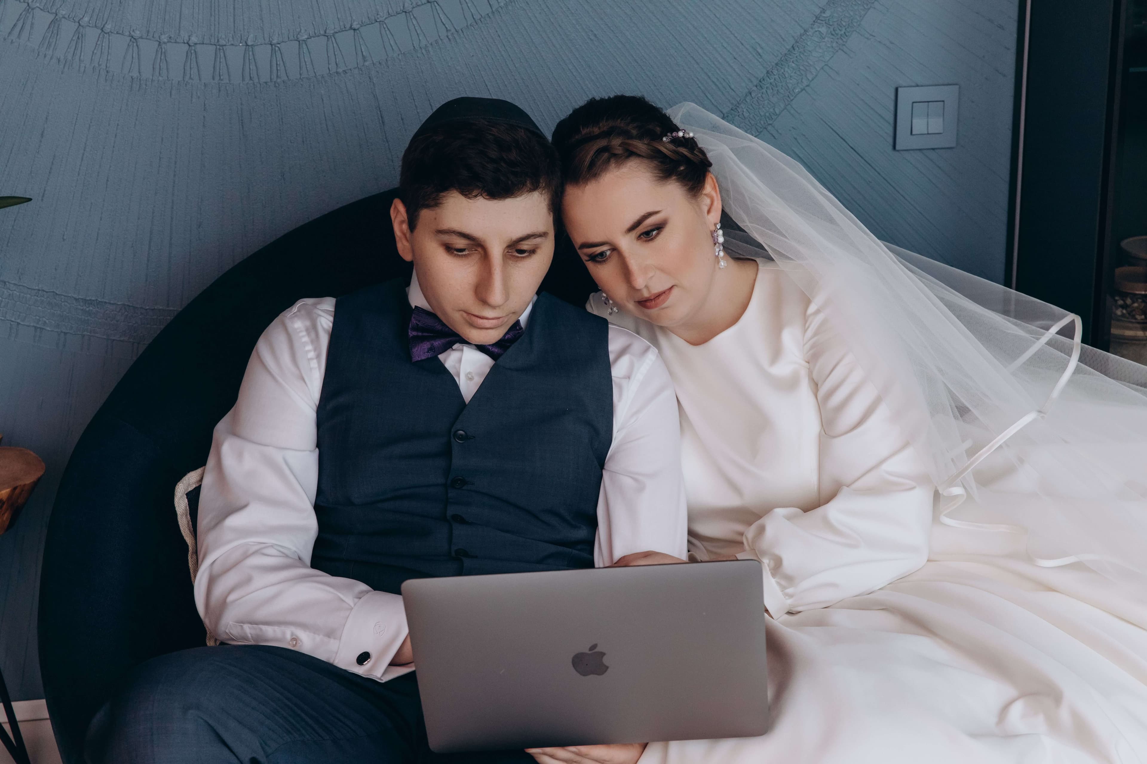 A newly married couple in wedding attire sitting together, looking at their wedding page on a laptop.